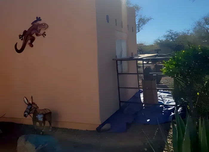 Peach stucco building exterior with late-afternoon light on a narrow patio with a small scaffold and tarp against the wall; metal gecko and donkey wall decorations, desert shrubs and agave at the right, clear blue sky beyond.