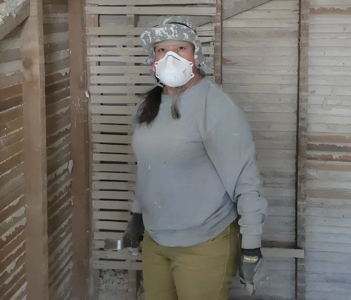 Partially demolished room with exposed wooden studs and lath with a Ross's Maintenance worker in a camouflage cap, N95 mask, grey sweatshirt, olive pants and gloves holds a short metal lath amid dust and debris as part of a Tucson home maintenance project.